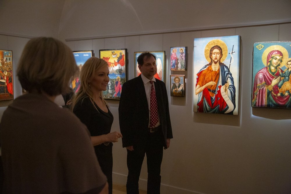 Trzy osoby stoją w przyćmionym wnętrzu galerii przed ścianą z oświetlonymi, kolorowymi ikonami religijnymi — centralnie mężczyzna w garniturze i krawacie, obok kobieta z blond włosami, po lewej sylwetka innej osoby tyłem, na ścianie m.in. duża ikona postaci z aureolą trzymającej zwój oraz ikona Matki Boskiej z Dzieciątkiem.