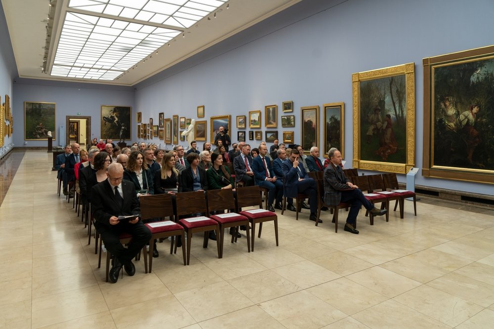 Grupa dorosłych osób w formalnych strojach siedzących w kilku rzędach drewnianych krzeseł z czerwonymi siedziskami w dużej sali galerii sztuki z jasnym świetlikiem sufitowym, marmurową posadzką, rzeźbą i licznymi obrazami w złoconych ramach na błękitnoszarych ścianach.