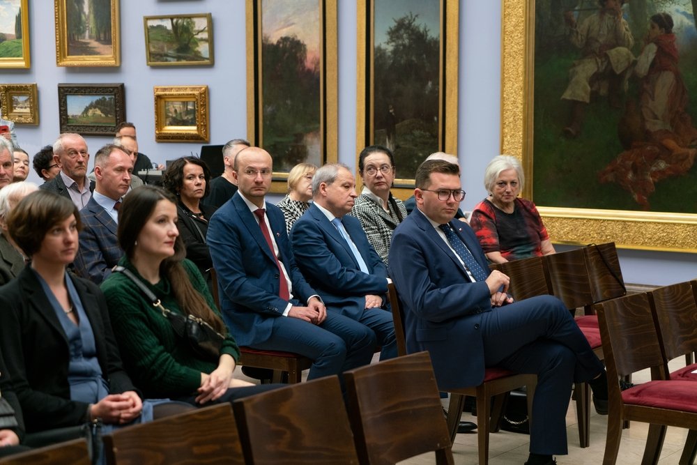 Grupa kilkunastu osób siedzi w rzędach drewnianych krzeseł w sali wystawowej z licznymi obrazami w złotych ramach na ścianach; na pierwszym planie kobiety w swetrach i eleganckich ubraniach oraz mężczyźni w garniturach, po prawej mężczyzna w granatowym garniturze i okularach siedzi z nogą założoną.