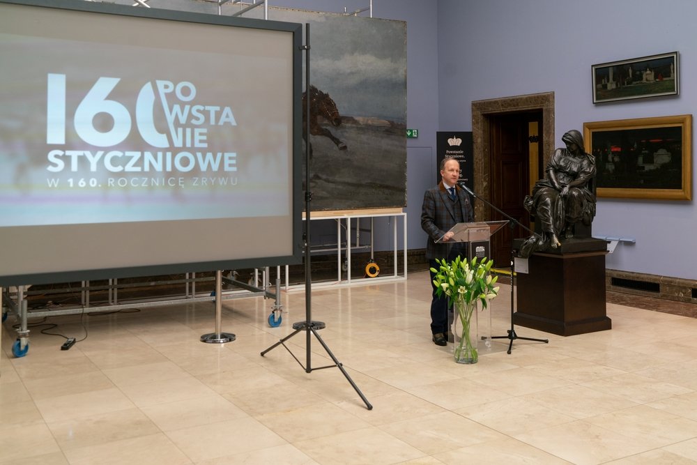 Galeria muzealna z dużym ekranem projekcyjnym po lewej z napisem „160 Powstanie Styczniowe”, mężczyzną przemawiającym przy przezroczystym pulpicie z mikrofonem pośrodku, wazonem z białymi liliami u stóp pulpitu oraz dużą brązową rzeźbą siedzącej postaci na cokole i obrazami wiszącymi na ścianie.
