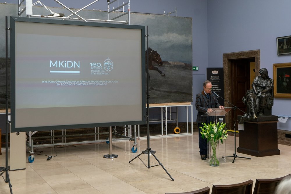 Wnętrze wystawowe z dużym ekranem projekcyjnym po lewej z widocznym napisem MKiDN i 160. rocznica, po prawej mężczyzna stoi przy przezroczystej mównicy z mikrofonem, przed nim wazon z białymi liliami, obok na cokole ciemna rzeźba siedzącej postaci, w tle duże malowidło konia i rusztowania.