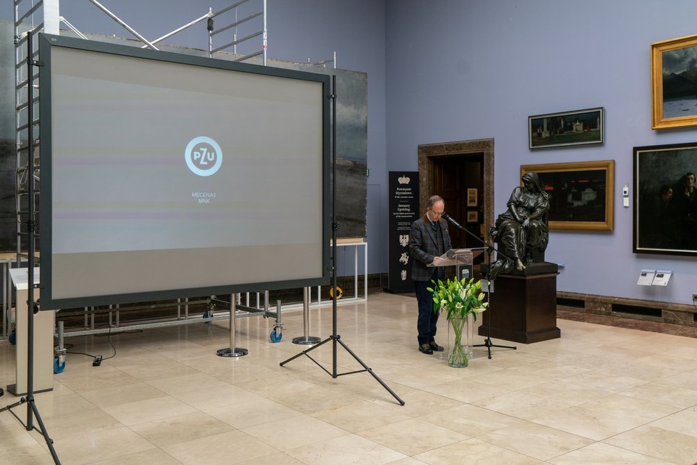 Wnętrze galerii z dużym ekranem projekcyjnym po lewej z widocznym logo PZU i rusztowaniem za nim, mężczyzną przy przezroczystej mównicy przemawiającym do mikrofonu, stojącą obok rzeźbą na cokole i wazonem z białymi liliami na jasnobeżowej, kafelkowej podłodze, w otoczeniu obrazów na jasnofioletowej ścianie.