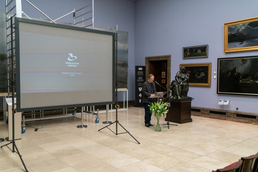 Duża sala muzealna z jasną, kafelkową posadzką i jasnofioletowymi ścianami; po lewej stoi duży ekran projekcyjny na statywie przed rusztowaniem, po prawej mężczyzna przy przezroczystej mównicy z mikrofonem obok ciemnej rzeźby na cokole, przed którą ustawiony jest wysoki szklany wazon z białymi liliami, a na ścianach wiszą obrazy w ozdobnych ramach, w prawym dolnym rogu widać rząd krzeseł.