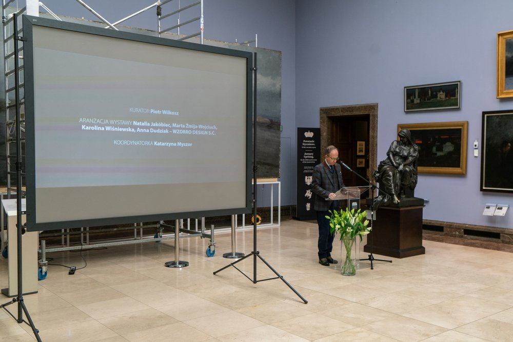Sala muzealna z dużym przenośnym ekranem projekcyjnym po lewej i rusztowaniem za nim, mężczyzną przy przezroczystym pulpicie z mikrofonem po prawej, stojącą obok rzeźbą na cokole oraz wazonem z białymi liliami, na ścianach kilka oprawionych obrazów.