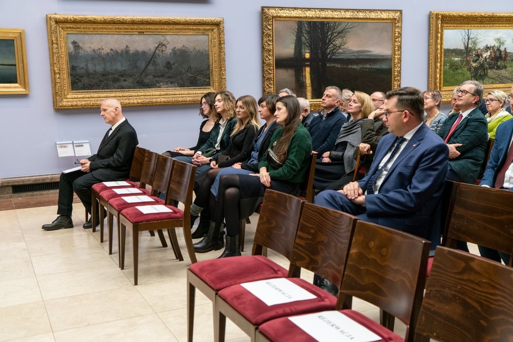 Kilkanaście osób siedzi w rzędach drewnianych krzeseł z bordowymi poduszkami w galerii z dużymi obrazami w złoconych ramach na ścianie; po lewej mężczyzna w czarnym garniturze coś zapisuje na kartce, a na kilku pustych siedzeniach leżą białe kartki.