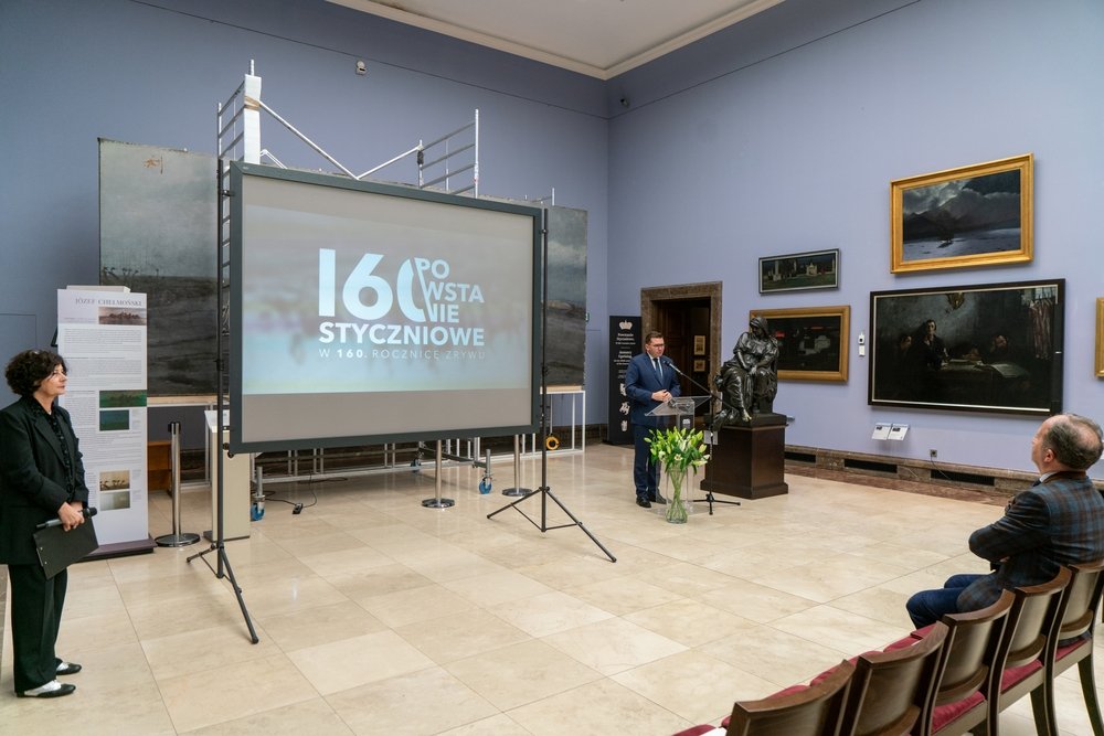 Sala wystawowa z fioletowymi ścianami i obrazami, na środku ekran projekcyjny z dużym napisem "160 Powstanie Styczniowe", obok stoi mównica z mężczyzną, przy niej rzeźba i wazon z tulipanami, z lewej kobieta trzymająca teczkę, a z prawej kilka rzędów krzeseł z jednym siedzącym mężczyzną.