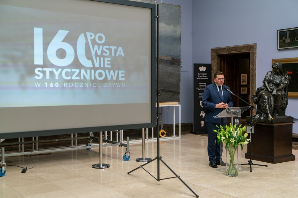 Mężczyzna w granatowym garniturze stoi przy przezroczystej mównicy z mikrofonami w sali wystawowej; po lewej duży ekran z napisem "160 Powstanie Styczniowe" i dopiskiem "w 160. rocznicę zrywu", po prawej rzeźba siedzącej kobiety na cokole i wazon z białymi liliami na podłodze.