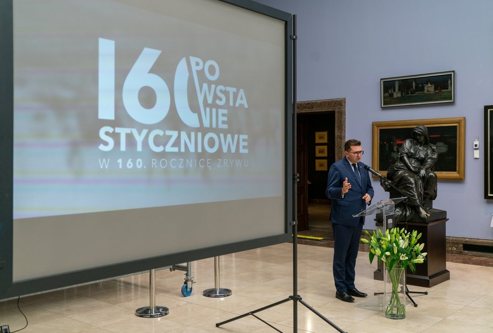 W sali wystawowej duży ekran projekcyjny po lewej z napisem "160 Powstanie Styczniowe", przy mównicy po prawej stoi mężczyzna w garniturze przemawiający do mikrofonu obok rzeźby siedzącej postaci na cokole, a przed mównicą stoi wazon z białymi tulipanami, w tle widoczne obrazy na ścianie.