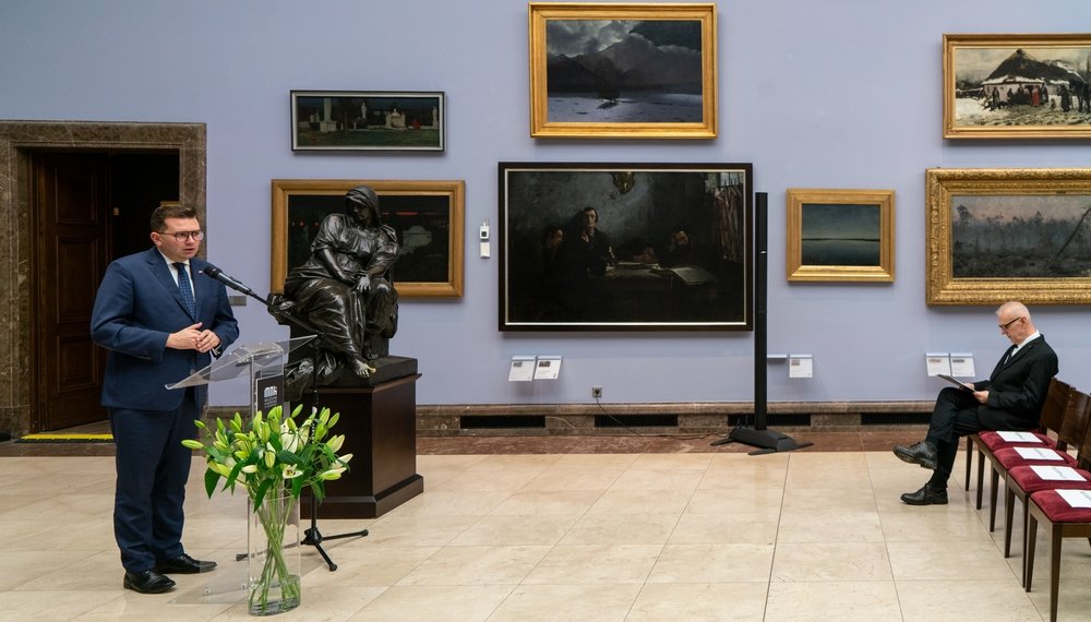 Wnętrze galerii z jasnofioletową ścianą wypełnioną oprawionymi obrazami; po lewej mężczyzna w granatowym garniturze przemawiający przy przezroczystym mównicy z mikrofonem, obok na cokole brązowa rzeźba siedzącej kobiecej postaci i wazon z białymi liliami, po prawej rząd czerwonych krzeseł, na jednym z nich siedzi mężczyzna w ciemnym garniturze czytający kartkę.