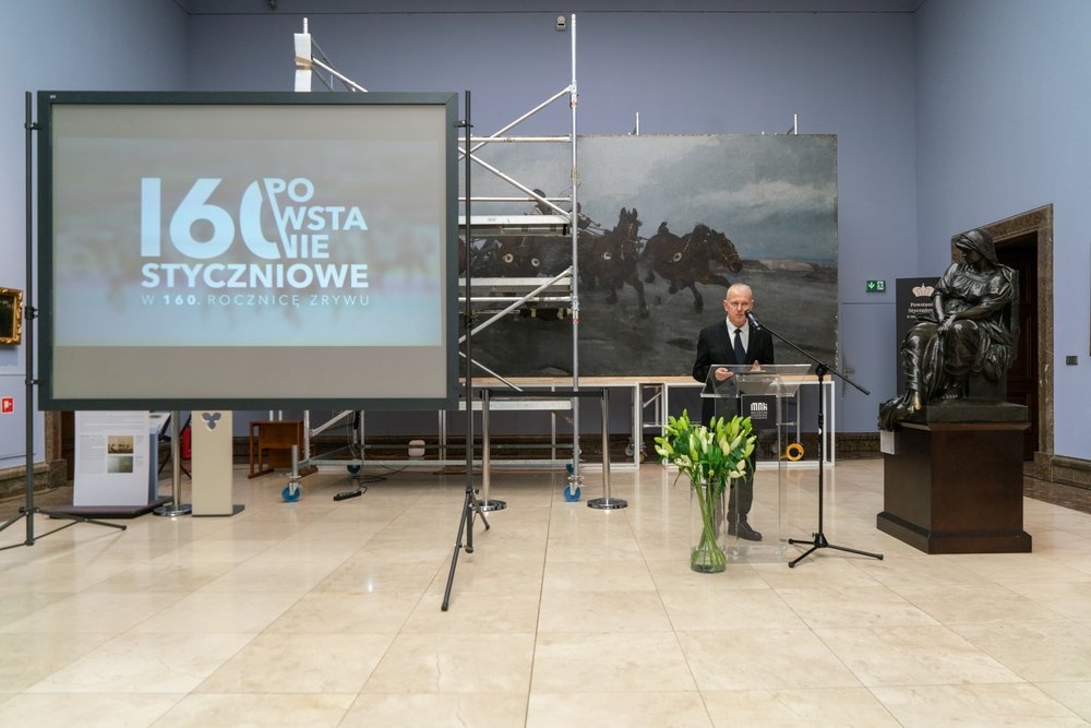 Sala muzealna o jasnoniebieskich ścianach i marmurowej podłodze z dużym ekranem projekcyjnym po lewej pokazującym napis "160 Powstanie Styczniowe", rusztowaniem i wielkim obrazem z końmi w tle, osobą w garniturze przemawiającą przy przezroczystym pulpicie z mikrofonem, wazonem z białymi liliami ustawionym przed pulpitem oraz brązowym posągiem siedzącej postaci na cokole po prawej.