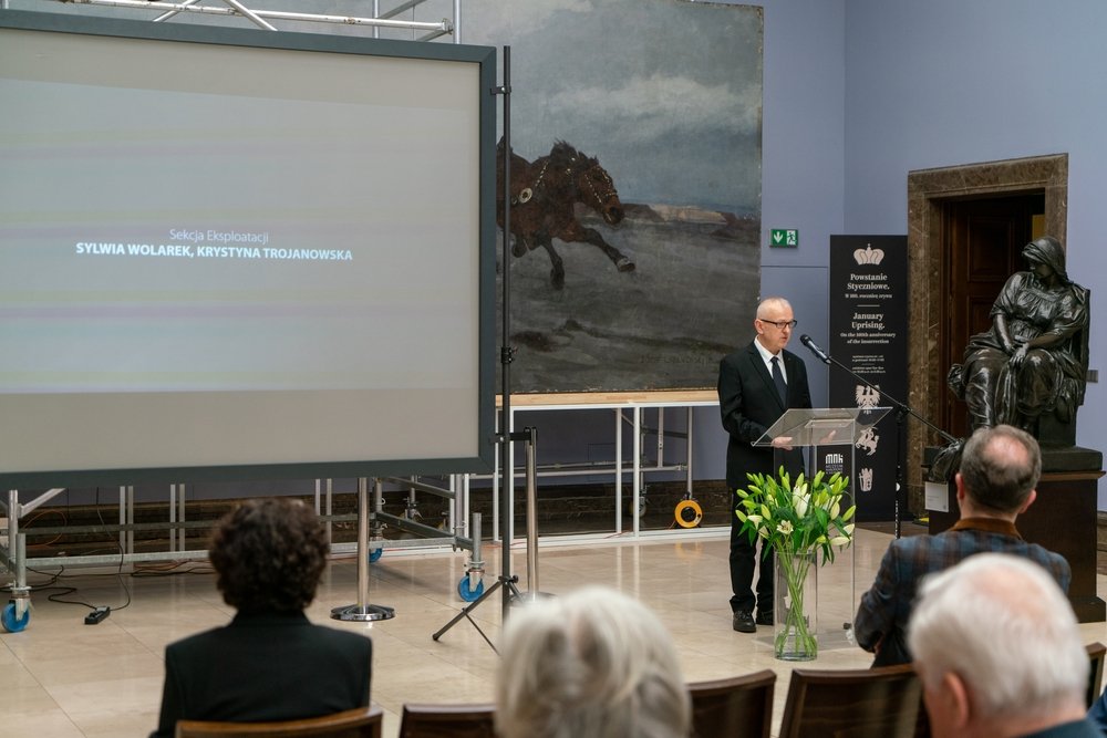 Mężczyzna w garniturze stoi przy przezroczystej mównicy z mikrofonem w sali wystawowej; po lewej duży ekran projekcyjny z napisem "SYLWIA WOLAREK, KRYSTYNA TROJANOWSKA", za nim wielkie płótno przedstawiające galopującego konia, po prawej rzeźba siedzącej postaci, przed mównicą wazon z białymi liliami, a w pierwszych rzędach kilka osób siedzi zwróconych przodem.