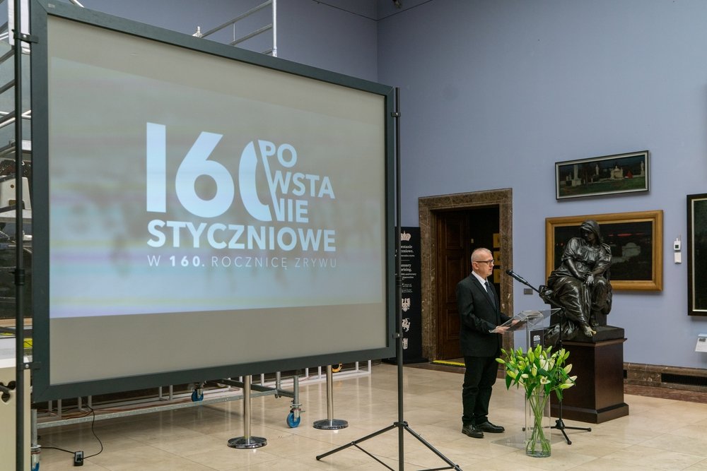 Wnętrze sali wystawowej z dużym ekranem po lewej wyświetlającym duży napis 160 oraz słowa STYCZNIOWE i mniejszy tekst W 160. ROCZNICĘ ZRYWU, po prawej mężczyzna w garniturze przemawiający przy szklanym pulpicie z mikrofonem, obok rzeźba siedzącej postaci na cokole, bukiet białych lilii w szklanym wazonie i wiszące obrazy na ścianie.