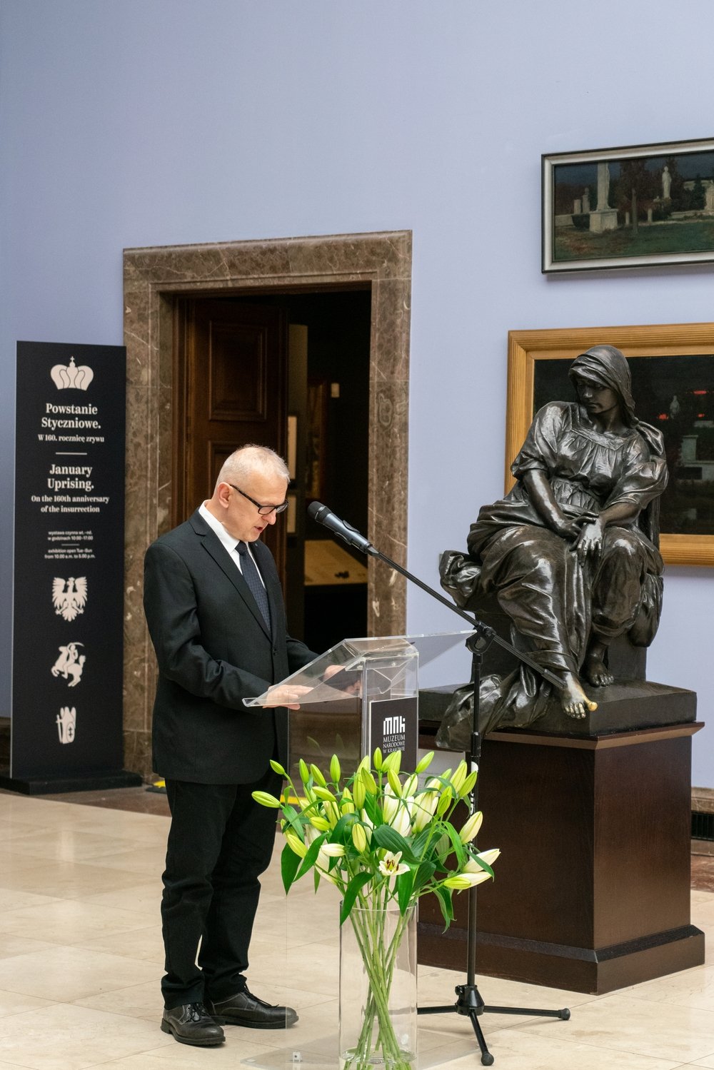 Mężczyzna w czarnym garniturze i okularach stoi przy przezroczystym pulpitu z mikrofonem czytając dokument, obok na cokole siedzi duża brązowa rzeźba kobiety, z przodu stoi wysoki szklany wazon z białymi liliami, a w tle widoczna jest tablica informacyjna o Powstaniu Styczniowym, otwarte drzwi i obrazy na jasnej ścianie sali muzealnej.
