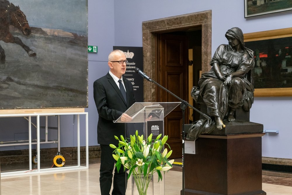 Mężczyzna w czarnym garniturze i okularach stoi za przezroczystym pulpitem i przemawia do mikrofonu w sali muzealnej; z przodu stoi wazon z białymi liliami, po prawej widoczna jest ciemna rzeźba siedzącej kobiety na cokole, a po lewej duży obraz na ścianie.