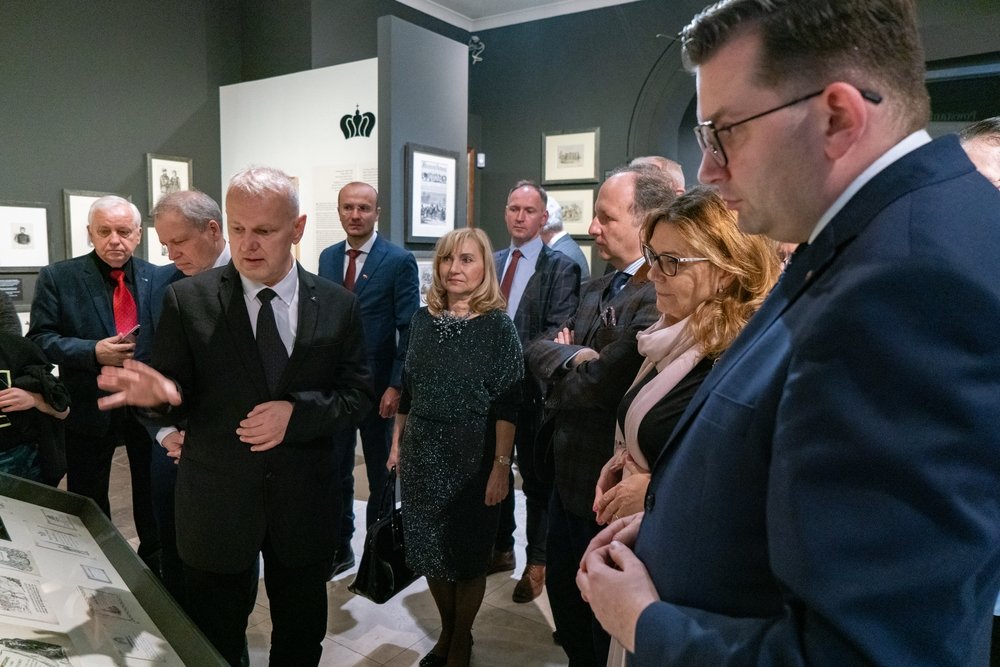 Grupa osób w eleganckich strojach stoi wokół przeszklonej gabloty w sali wystawowej z oprawionymi rysunkami na ścianach; mężczyzna w ciemnym garniturze gestykuluje nad gablotą, obok kobieta w ciemnej sukience trzyma torebkę, a po prawej kilka osób w marynarkach przygląda się eksponatom.