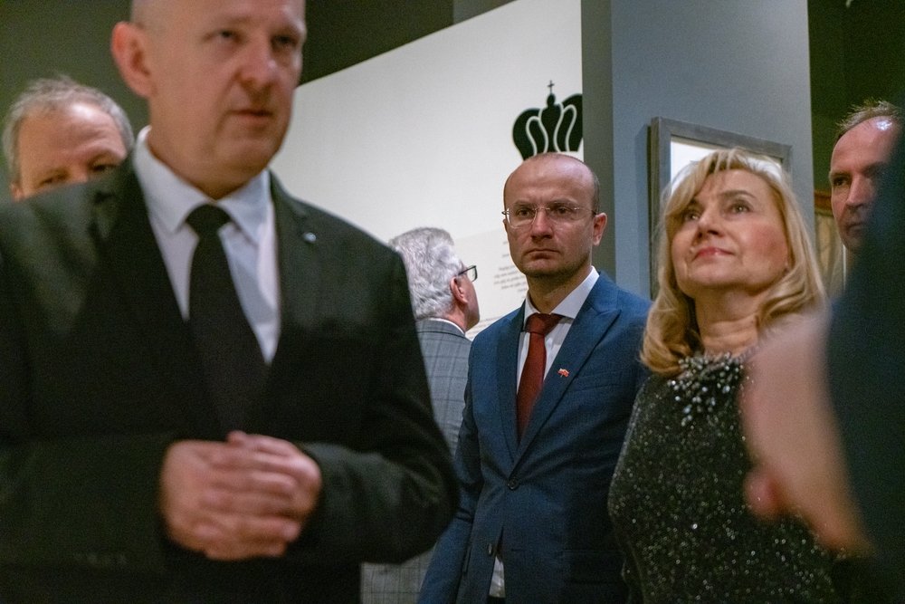 Kilka formalnie ubranych osób w pomieszczeniu z obrazami: na środku łysy mężczyzna w okularach, granatowym garniturze i czerwonym krawacie, po prawej blond kobieta w błyszczącej czarnej sukience patrząca w górę, z przodu częściowo rozmyty mężczyzna w czarnym garniturze ze złożonymi dłońmi, w tle fragment ramy obrazu i czarny emblemat korony na ścianie.