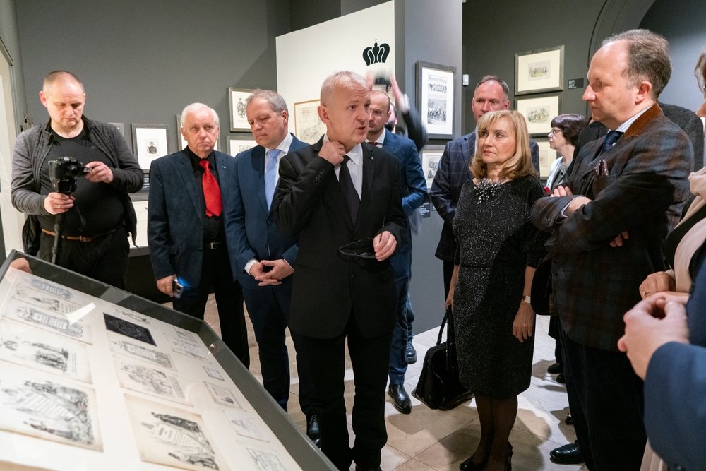 Grupa formalnie ubranych osób stoi w sali wystawowej przy pochylonej gablocie z rysunkami i grafikami; w centrum mężczyzna w garniturze trzyma okulary i poprawia kołnierz, obok stoi kobieta w czarnej sukience z torebką, po lewej mężczyzna z aparatem, a na ścianach w tle wiszą oprawione prace.