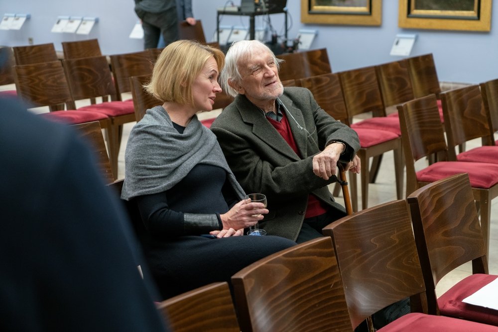 Starszy mężczyzna z białymi włosami i laską siedzi obok kobiety o krótkich blond włosach i szarym szalu, która trzyma kieliszek, oboje na rzędach drewnianych krzeseł z czerwonymi poduszkami w sali z obrazami na ścianach.