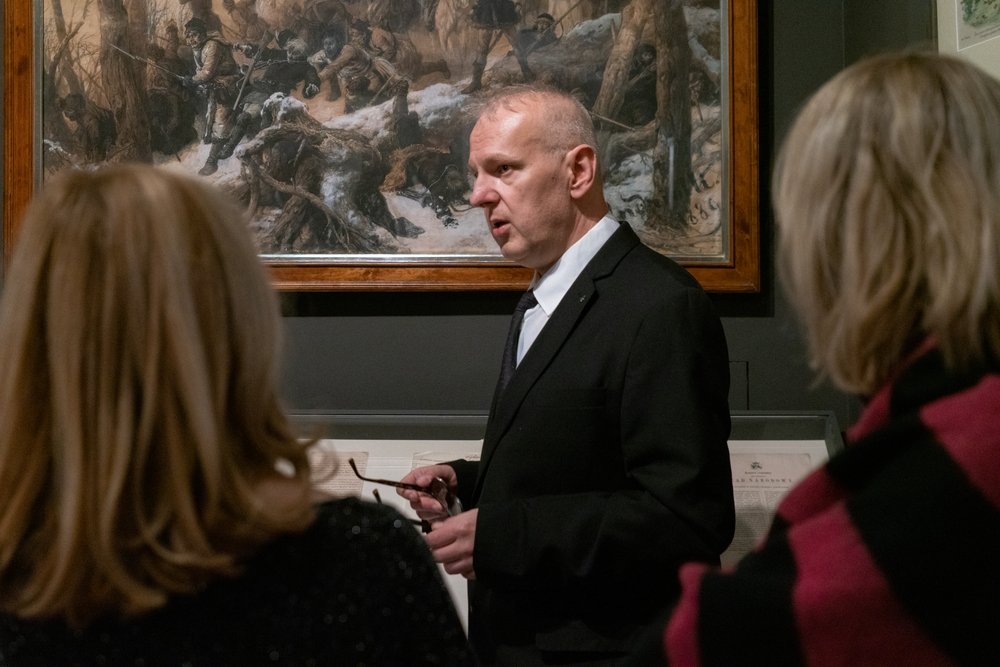 Mężczyzna w czarnym garniturze stoi bokiem przed oprawionym obrazem w galerii, trzymając okulary, a z tyłu na pierwszym planie widać dwie kobiety zwrócone ku niemu.