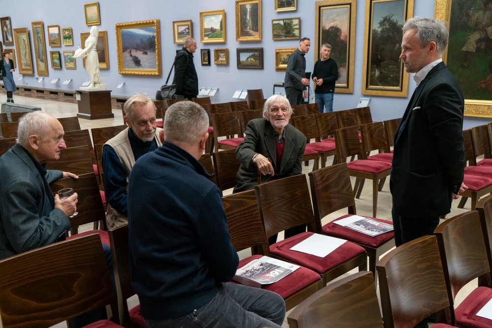 Kilku starszych mężczyzn siedzi w rzędach drewnianych krzeseł z czerwonymi poduszkami w galerii sztuki, obok stoi mężczyzna w garniturze; wokół wiszą liczne oprawione obrazy, na niektórych krzesłach leżą gazetki i kartki, a w tle widać białą rzeźbę i innych zwiedzających.