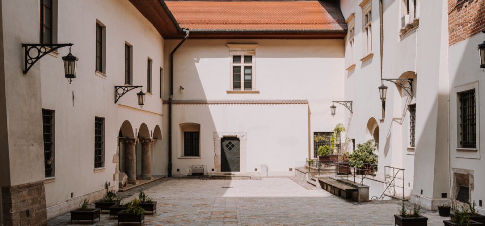 Brukowany, pusty dziedziniec otoczony jasnymi budynkami z czerwonym dachem, z arkadowym przejściem i kamiennymi kolumnami po lewej, oknami z kratami, pojedynczymi drzwiami w środku ściany, kutymi lampami ściennymi oraz donicami z roślinami i ławkami przy krawędziach.