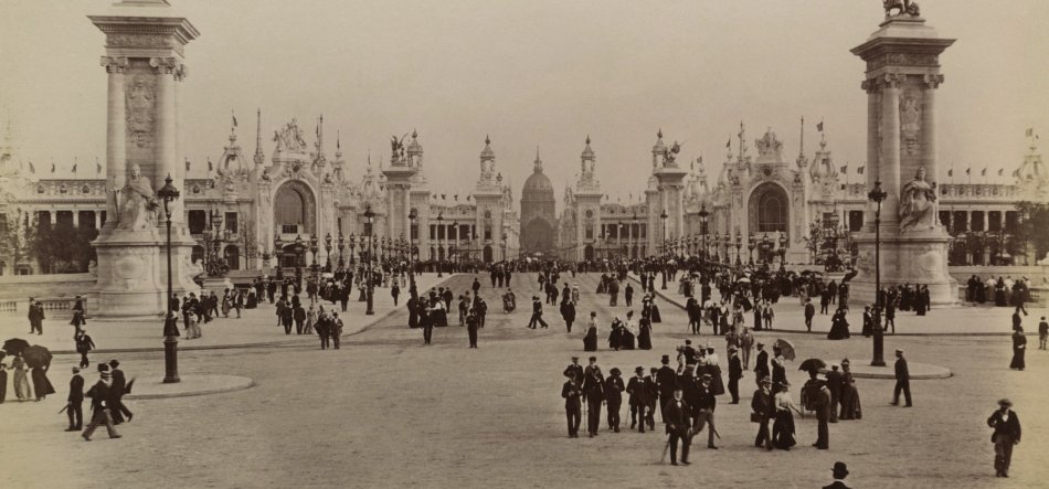 Sepiafotografia przedstawiająca szeroki brukowany plac i środkową aleję prowadzącą do rozległego, ozdobnego gmachu z centralną kopułą, arkadami i wieżami, po bokach dwie wysokie kolumny z rzeźbami, liczne latarnie i flagi oraz tłumy postaci w historycznych strojach rozchodzących się i stojących na placu.