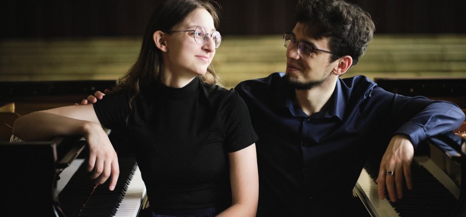 Kobieta i mężczyzna siedzą obok siebie przy dwóch fortepianach — kobieta w czarnym topie ma jedną rękę na klawiaturze, mężczyzna w ciemnej koszuli opiera dłonie na drugim instrumencie i ma rękę na ramieniu kobiety, oboje w okularach, w tle rozmyte drewniane ławki.