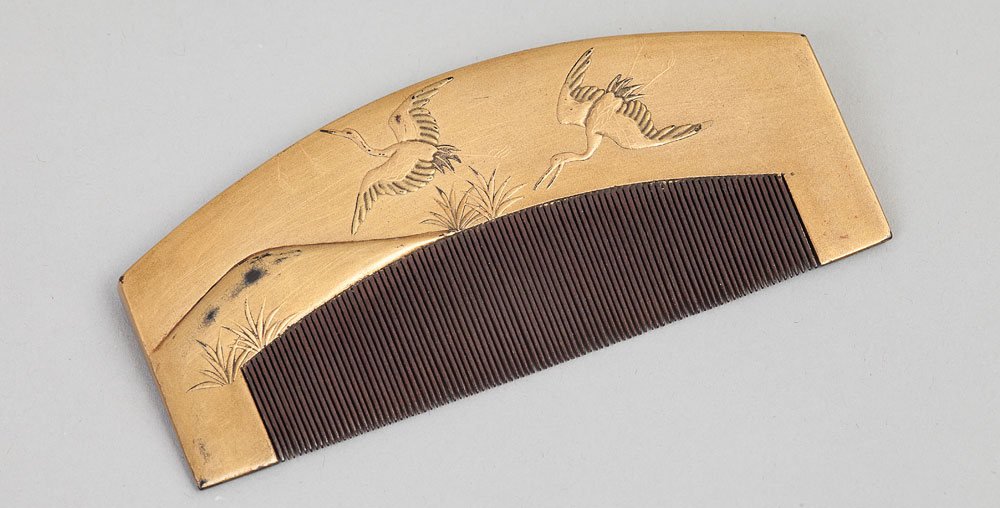 Poziomo ułożony ozdobny grzebień z gęstymi ciemnobrązowymi zębami i złotą metalową górną częścią zdobioną rytowanymi dwoma lecącymi ptakami i trawą na jasnoszarym tle.