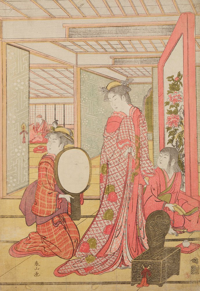 Japoński drzeworyt przedstawiający wnętrze z trzema kobietami w kimono — centralna stojąca postać w długim czerwono‑różowym, wzorzystym kimonie, z przodu klęczy kobieta trzymająca okrągłe lusterko, po prawej siedzi druga kobieta w czerwonym stroju; na podłodze wzorzyste tkaniny, po prawej ozdobny parawan z motywem kwiatowym i zdobna skrzynia, w głębi przesuwane panele i dwie postacie w sąsiednim pomieszczeniu.