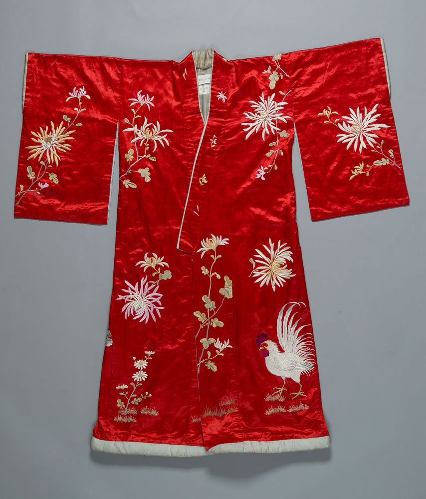 Czerwone jedwabne kimono rozłożone na płasko na jasnoszarym tle, z szerokimi rękawami i białą lamówką u dołu, ozdobione haftami złoto-białych chryzantem, pnączy i liści oraz dużym haftowanym białym kogutem z czerwonymi i fioletowymi akcentami.