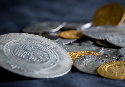 Rozrzucone metalowe monety w różnych rozmiarach i kolorach — duża srebrna moneta z wyraźnym wytłoczonym wzorem na pierwszym planie oraz mniejsze srebrne i złote monety na ciemnym materiale, z płytką głębią ostrości.