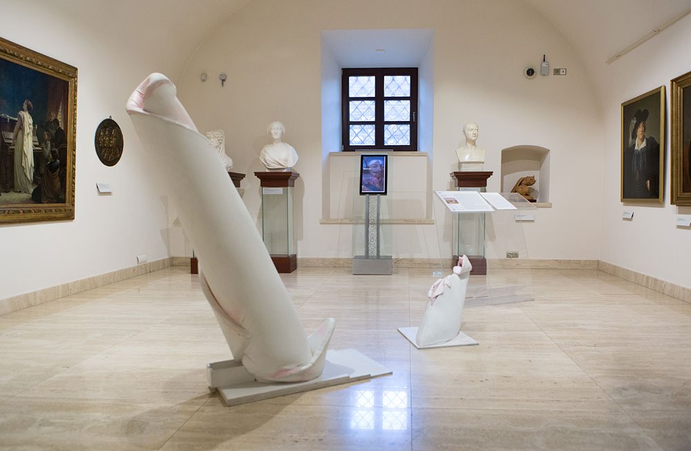 Widok galerii muzealnej z jasną marmurową podłogą i łukowatym sufitem, w centrum dwa abstrakcyjne białe rzeźby o wydłużonych, zaokrąglonych kształtach z różowymi akcentami ustawione na podstawkach, wokół nich popiersia na kolumnach, tablice opisowe oraz oprawione obrazy na ścianach, a w tle małe okno z kratownicą.