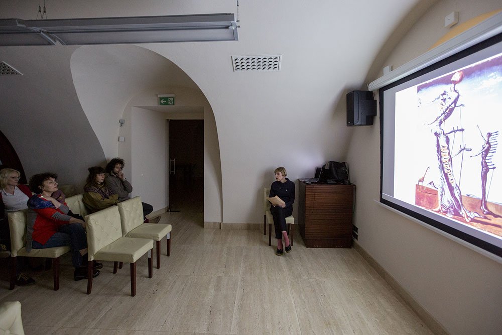 Wnętrze małej sali z zaokrąglonym sklepieniem: po prawej duży ekran z projekcją obrazu przedstawiającego wydłużone figury, obok szafka z głośnikiem i siedząca osoba trzymająca kartki, po lewej rząd kremowych krzeseł z kilkoma osobami zwróconymi w stronę ekranu, w tle ciemne przejście i znak wyjścia ewakuacyjnego.