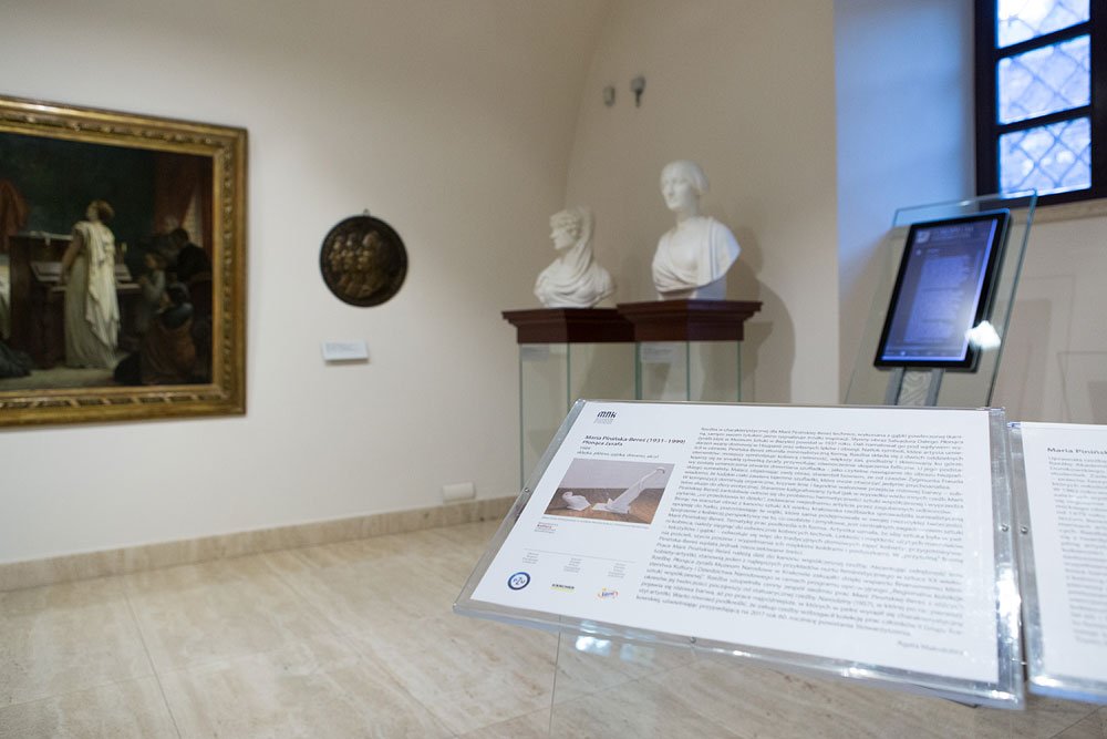 Wnętrze galerii: na pierwszym planie informacyjna plansza z tekstem i małym zdjęciem w przezroczystym stojaku, w tle po prawej dwa białe popiersia na drewnianych postumentach oraz stojak z dotykowym ekranem przy oknie, a po lewej duży oprawiony obraz i okrągła dekoracyjna płaskorzeźba na jasnej ścianie nad jasną posadzką.
