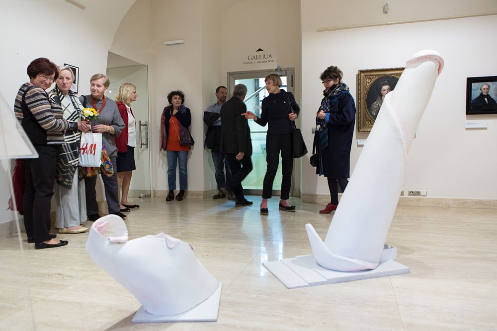 Wnętrze galerii sztuki z grupą osób stojących po lewej i przy drzwiach z napisem "GALERIA", na podłodze dwie duże białe abstrakcyjne rzeźby, na ścianach oprawione portrety.