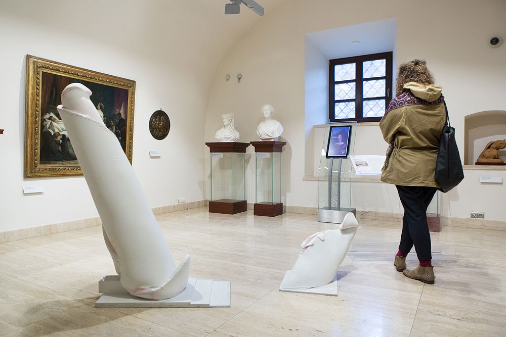 Wnętrze galerii z dwiema białymi abstrakcyjnymi rzeźbami na podłodze na pierwszym planie, po lewej obraz w złotej ramie, w głębi dwa marmurowe popiersia na szklanych podstawach i przy oknie stojący ekran informacyjny, a zwrócona tyłem zwiedzająca w zielonej kurtce z czarną torbą ogląda ekspozycję.