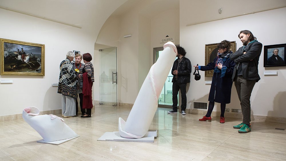 Wnętrze jasnej galerii ze sklepionym sufitem i jasną marmurową podłogą, na środku dwie białe abstrakcyjne rzeźby o wydłużonych zaokrąglonych kształtach z różowymi akcentami ustawione na podstawach; po lewej trzy osoby rozmawiają przy obrazie w złotej ramie, po prawej trzy osoby stoją i oglądają prace wiszące na ścianie, w głębi przeszklone drzwi z napisem GALERIA.