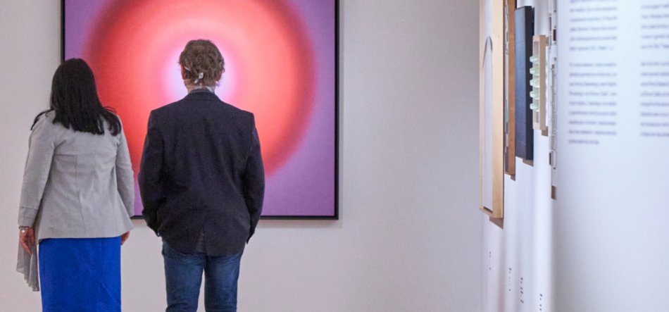 Dwie osoby widziane od tyłu stoją przed dużym oprawionym obrazem z koncentrycznym czerwono‑różowym okręgiem na białej ścianie galerii; po lewej kobieta w jasnej marynarce i niebieskiej spódnicy, po prawej mężczyzna w ciemnej marynarce i dżinsach, dalej po prawej widoczne kolejne prace i panel z tekstem.
