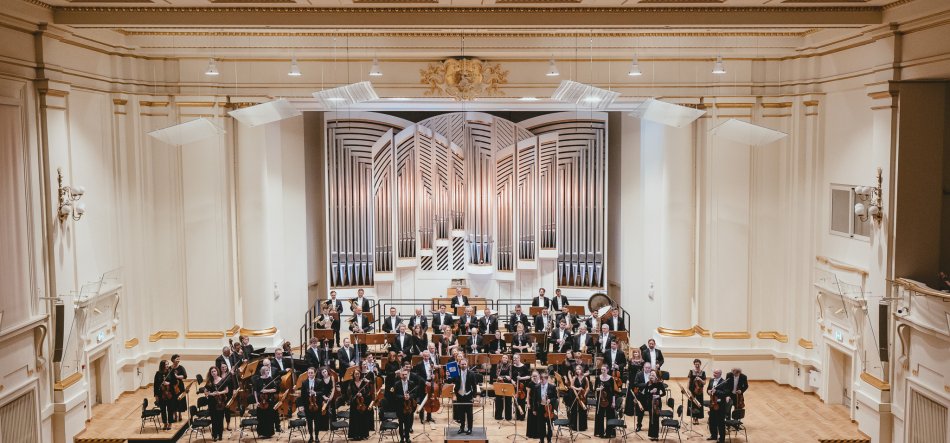 Pełna orkiestra symfoniczna ustawiona na drewnianej scenie dużej, jasnej sali koncertowej z ozdobnymi ścianami i złotymi akcentami, na tle wysokiego piszczałowego organu; na scenie szeregi muzyków z instrumentami smyczkowymi, dętymi i perkusją, a z przodu stoi dyrygent.