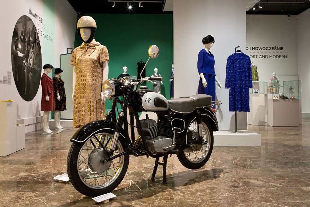 Czarny zabytkowy motocykl na podstawie ustawiony na marmurowej posadzce w sali wystawowej, w tle manekiny w sukienkach — jeden w żółto-brązowej sukience z kaskiem, drugi w niebieskim komplecie oraz wisząca niebieska sukienka, gabloty ekspozycyjne i kolorowe ściany muzeum.