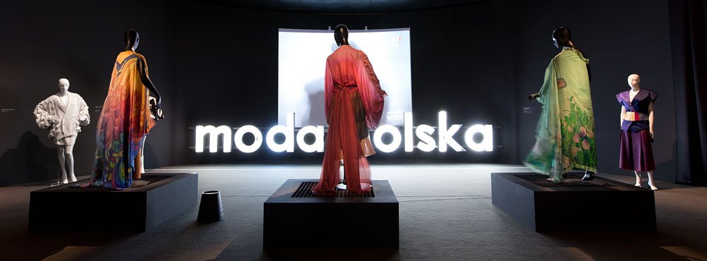 Ciemna sala wystawowa z pięcioma manekinami ustawionymi na podestach w kolorowych, drapowanych strojach oraz centralnie umieszczonym podświetlonym białym napisem moda polska