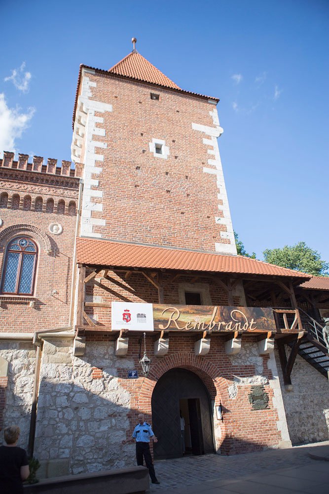 Wysoka ceglana wieża z czerwoną dachówką i jasnymi krawędziami, kamiennym parterem i drewnianym balkonem z dużym szyldem "Rembrandt" nad łukowym wejściem, obok którego stoi mężczyzna w jasnoniebieskim mundurze; po lewej ozdobne okno, po prawej zewnętrzne drewniane schody, a nad wszystkim błękitne niebo.