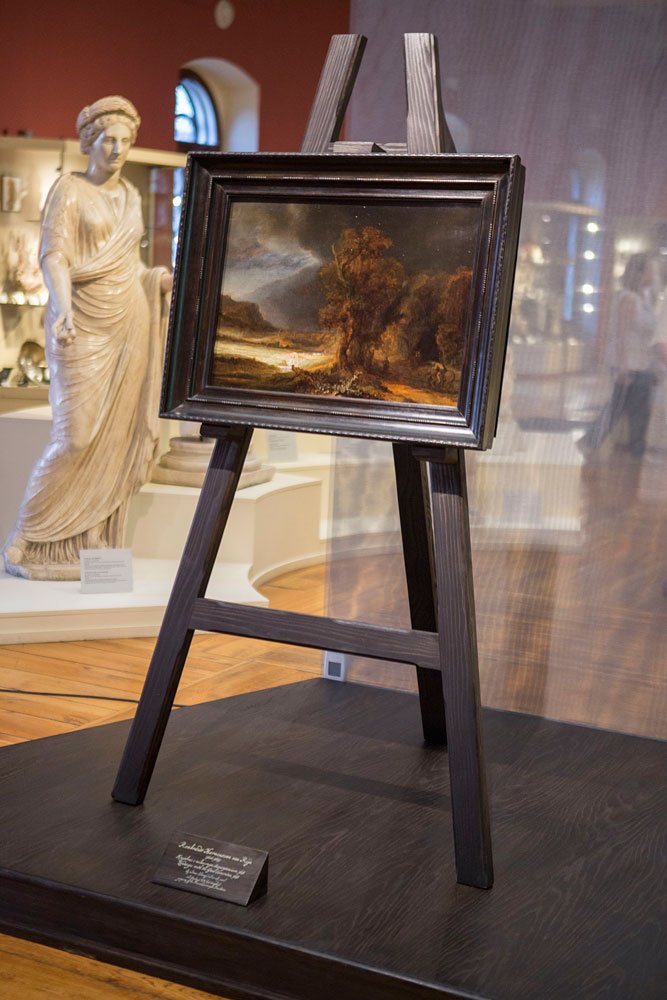 Na drewnianej platformie w muzeum stoi ciemny sztalugowy stojak z oprawionym obrazem pejzażu, obok po lewej widoczny marmurowy posąg kobiecy w drapowanej szacie, w tle gabloty z eksponatami, łukowate okno, ciepłe oświetlenie i drewniana podłoga, a z przodu leży mała tabliczka ekspozycyjna.