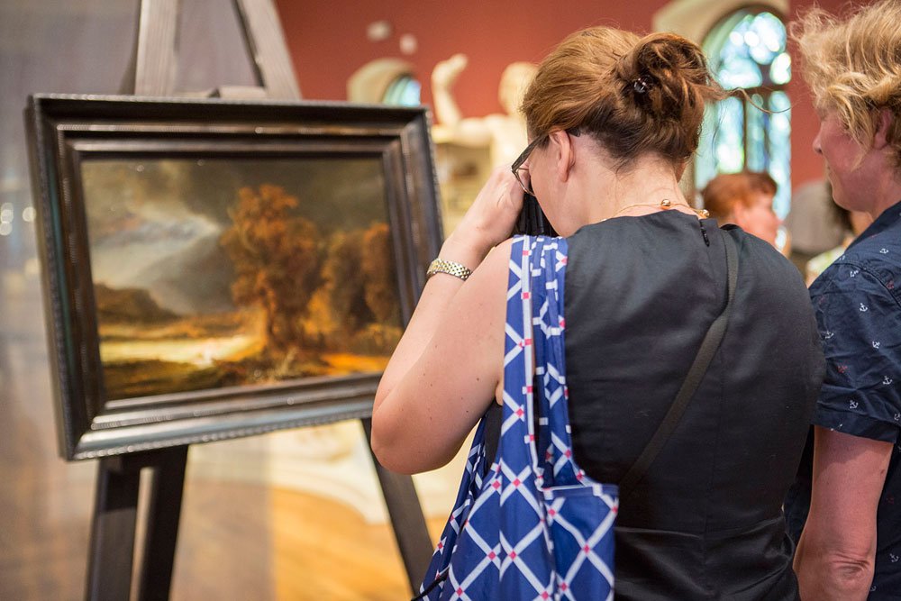 Kobieta widziana od tyłu robi zdjęcie obrazowi pejzażowemu na sztaludze w galerii; ma upięte włosy, czarną sukienkę i niebieską torbę z geometrycznym wzorem, obok stoi inna osoba, w tle rzeźby, czerwone ściany i łukowate okno.