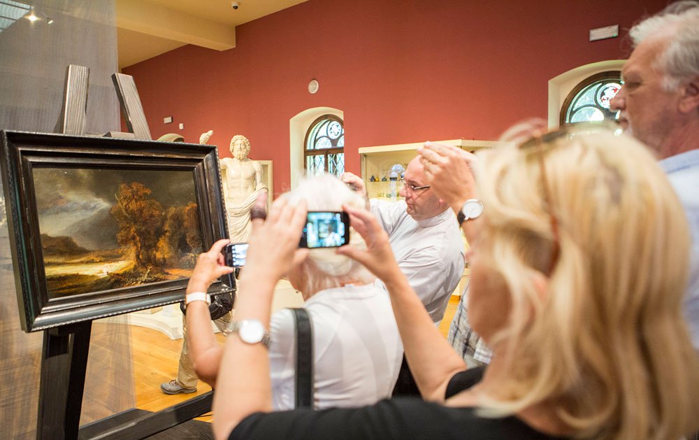 Grupa zwiedzających w muzeum — rozmyte głowy i uniesione dłonie z telefonami fotografujące olejny pejzaż w czarnej ramie na sztaludze, za którym stoi marmurowa rzeźba postaci, czerwone ściany z łukowymi oknami i drewniana podłoga.