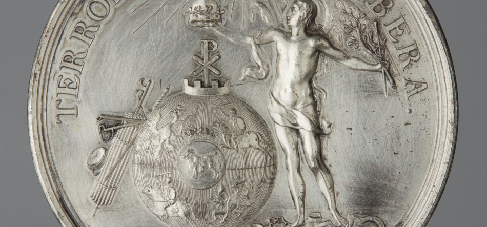Srebrny medal w zbliżeniu z reliefem przedstawiającym po prawej nagą męską postać przepasaną u bioder, trzymającą gałąź i opierającą rękę o wypukłą kulę ziemską z widoczną mapą, koroną i symbolem Chi‑Rho na szczycie, po lewej wiązka rózg z toporem (fasces), częściowy napis na obwodzie "TERR..." i "BERA" oraz widoczne rysy i faktura metalu.