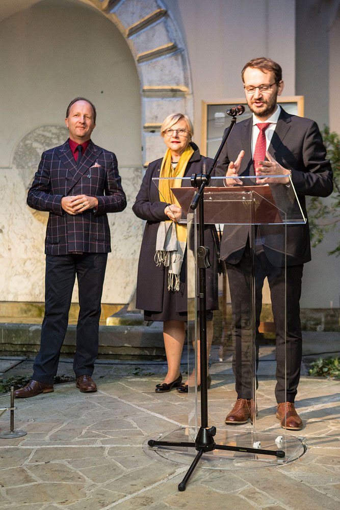 Trzech dorosłych stoi na kamiennym podłożu przy przezroczystym pulpicie z mikrofonem: z przodu po prawej mężczyzna w ciemnym garniturze, czerwonym krawacie i okularach gestykuluje przy mównicy, za nim kobieta w ciemnym płaszczu z żółtym szalem, a po lewej mężczyzna w kraciastej marynarce i czerwonej koszuli; w tle marmurowa ściana i kamienny łuk.