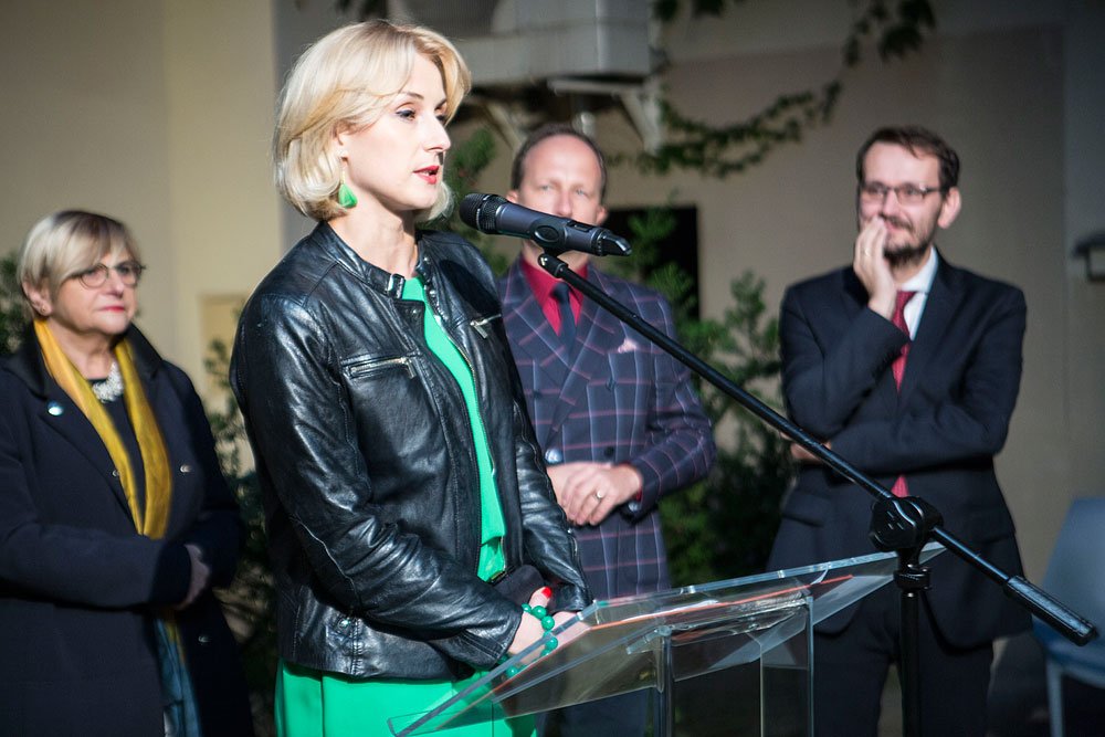 Kobieta o krótkich blond włosach w zielonej sukience i czarnej skórzanej kurtce stoi przy mikrofonie za przezroczystą mównicą, a za nią stoją trzy osoby: z lewej kobieta w okularach z żółtym szalikiem, na środku mężczyzna w kraciastej marynarce, a z prawej mężczyzna z brodą i okularami trzymający dłoń przy twarzy.