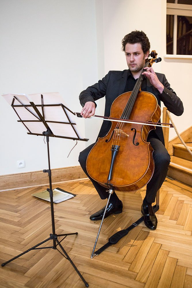 Mężczyzna w ciemnym garniturze siedzi na krześle i gra na wiolonczeli trzymając smyczek na strunach, przed nim stoi statyw z nutami, instrument oparty końcówką na drewnianym parkiecie, w tle schody i okno.