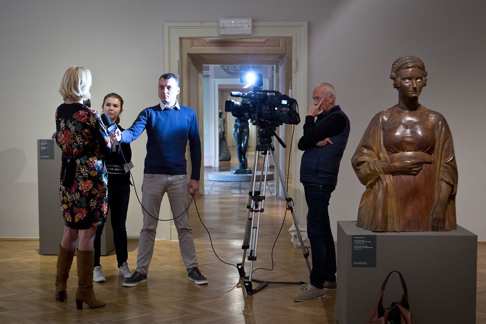 Wnętrze galerii sztuki: po lewej kobieta w kwiecistej sukience udzielająca wywiadu mężczyźnie w niebieskim swetrze trzymającemu mikrofon i młodszej kobiecie z kablami, na środku kamera na statywie z włączonym światłem i stojący obok operator z ręką przy twarzy, po prawej duża drewniana rzeźba kobiety na postumencie, w głębi przez otwarte drzwi widoczne kolejne rzeźby i drewniana podłoga.